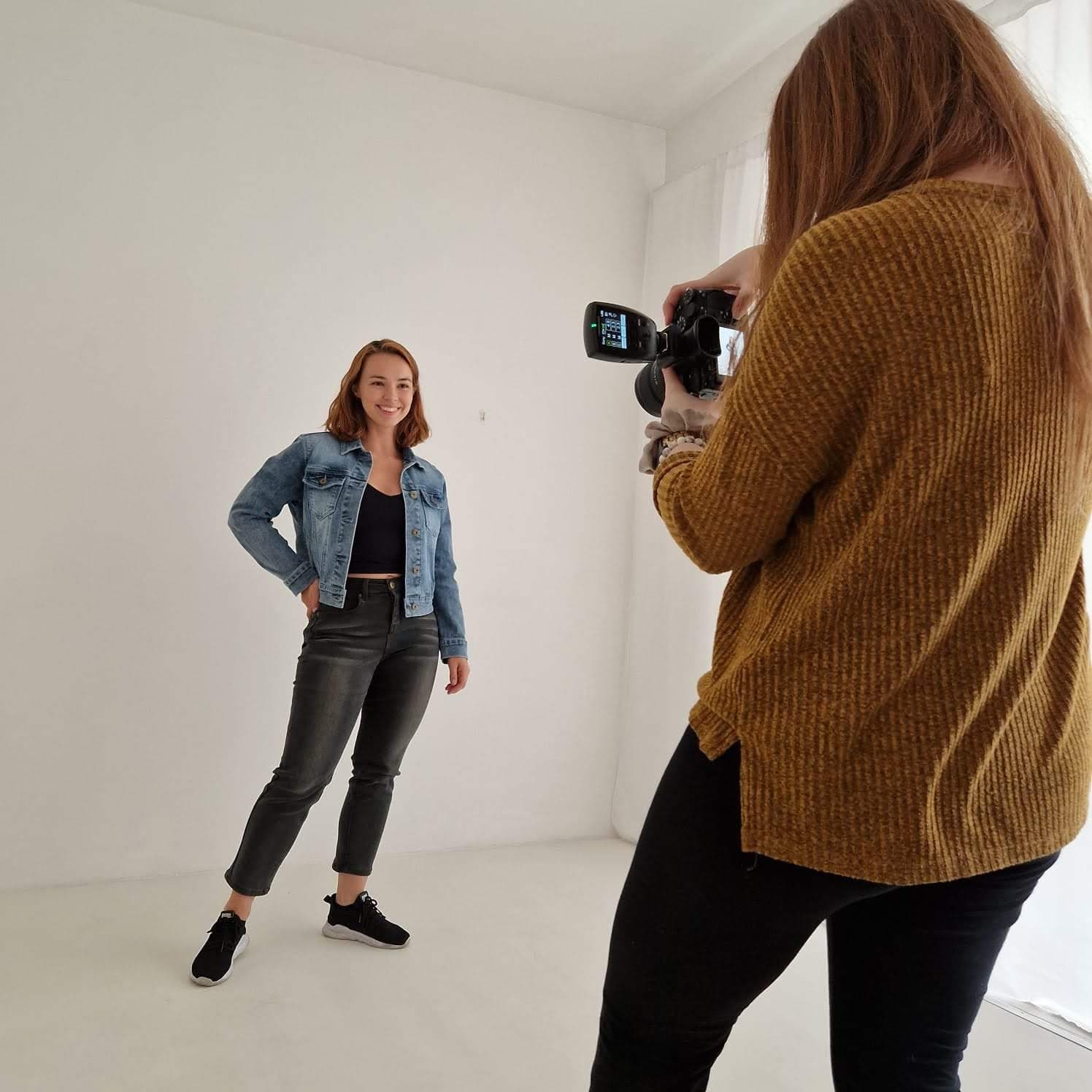 Lucy modelling a denim jacket and black jeans in a white photo studio while a photographer takes pictures